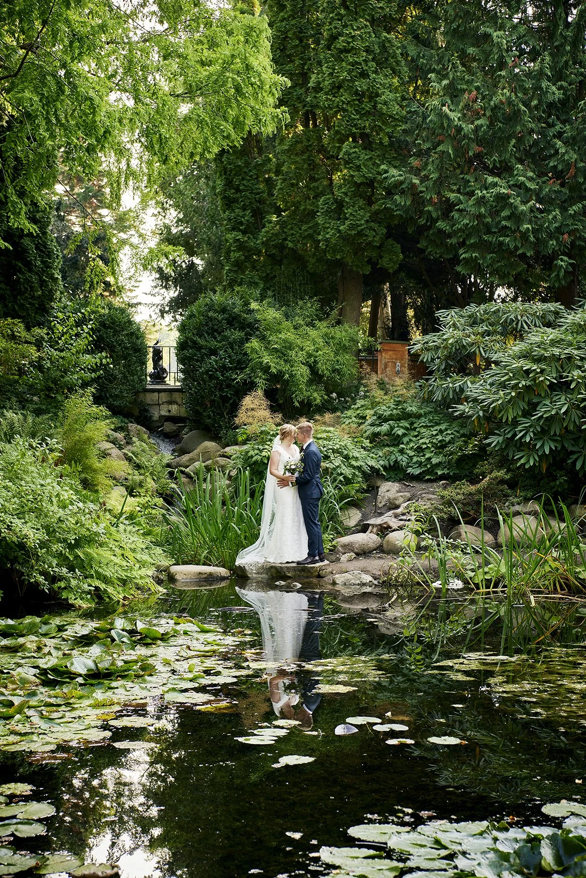 A bride and groom standing on rocks in a pond, surrounded by lush green trees and plants, with their reflection visible in the water.