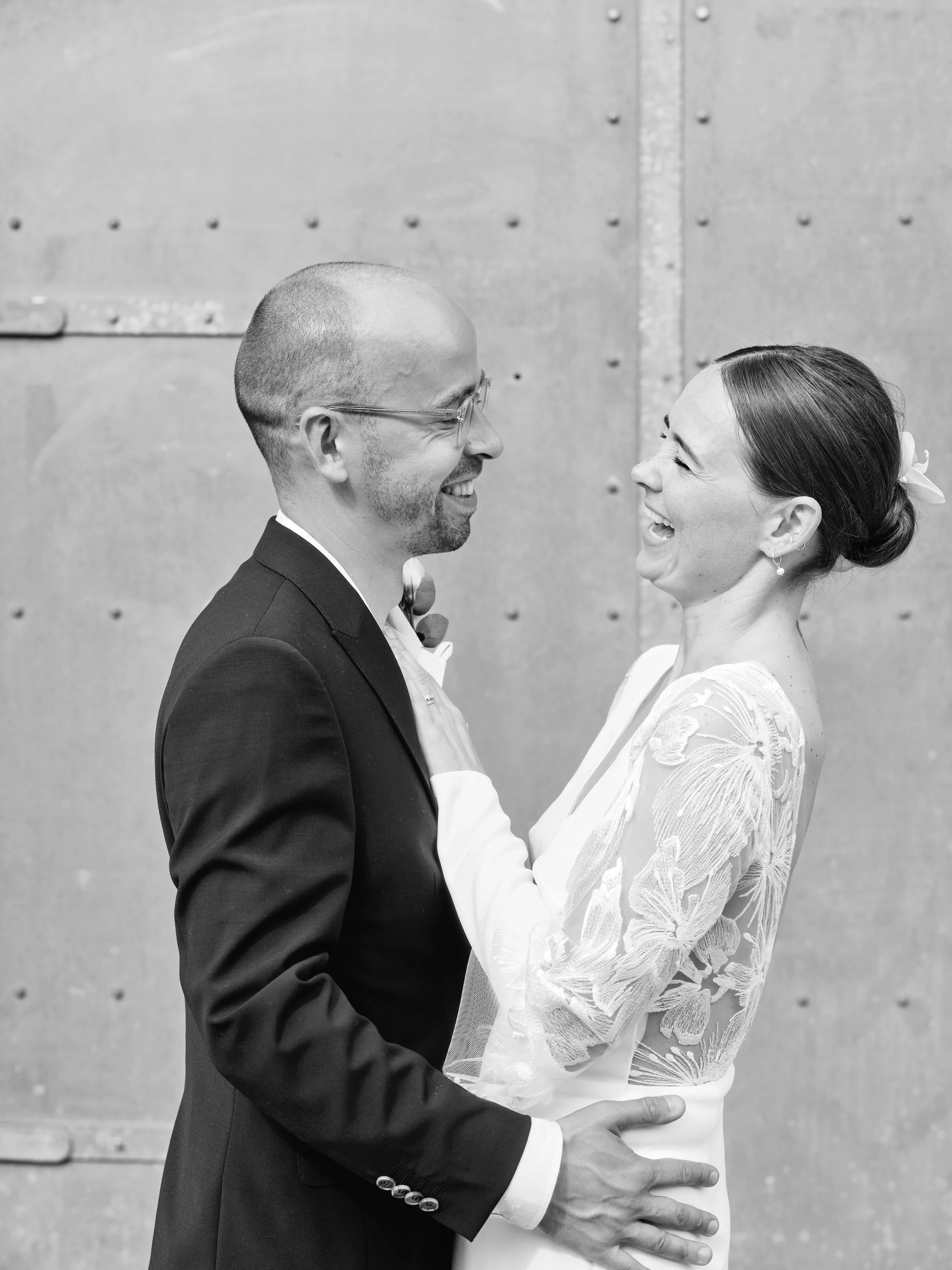 A black and white photo of a happy bride and groom gazing at each other, holding hands, standing in front of a textured wall.