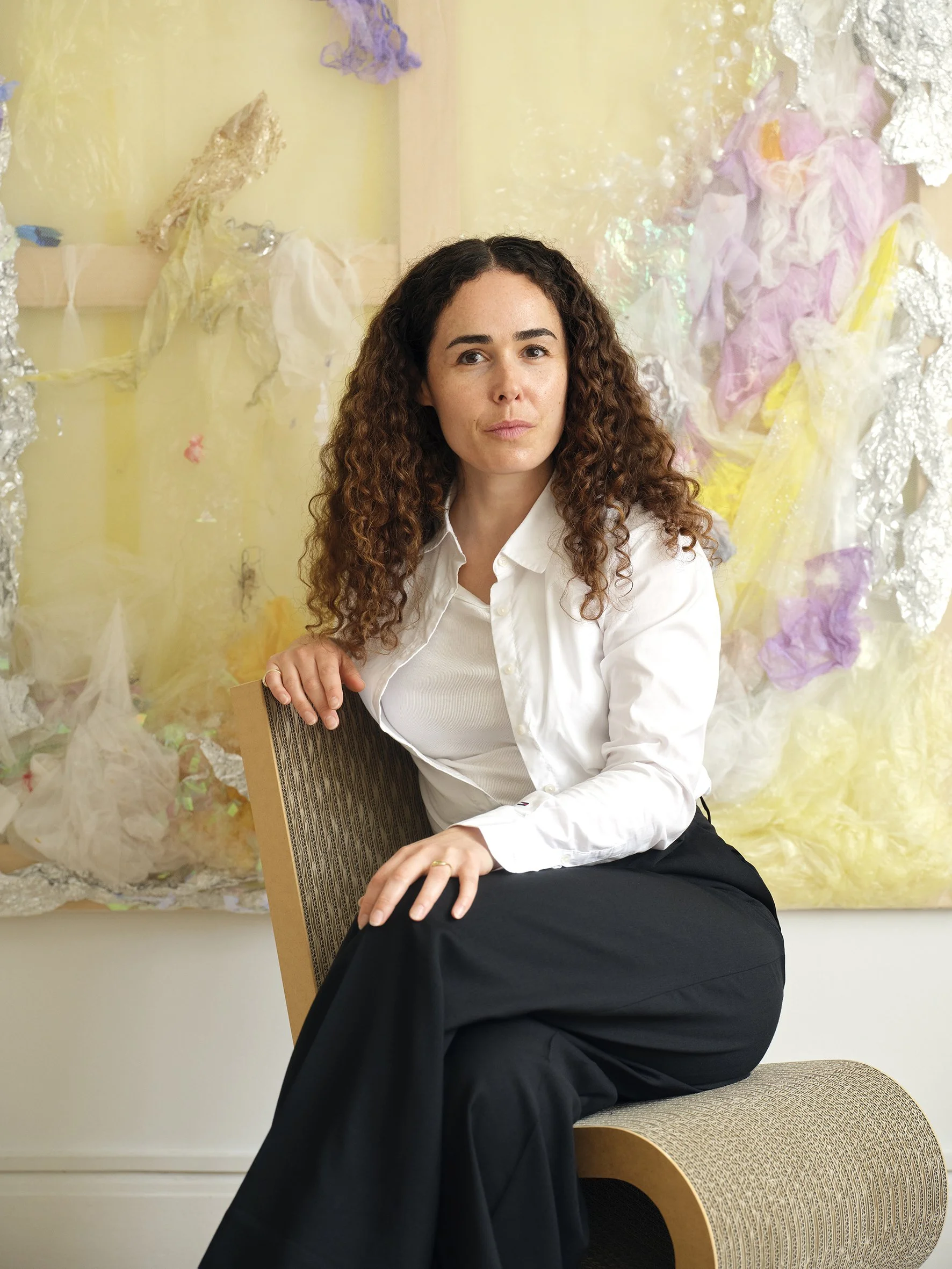 A woman with curly brown hair dressed in a white shirt and black pants sitting on a beige chair in front of a colorful abstract background. Laura curator and art collector.