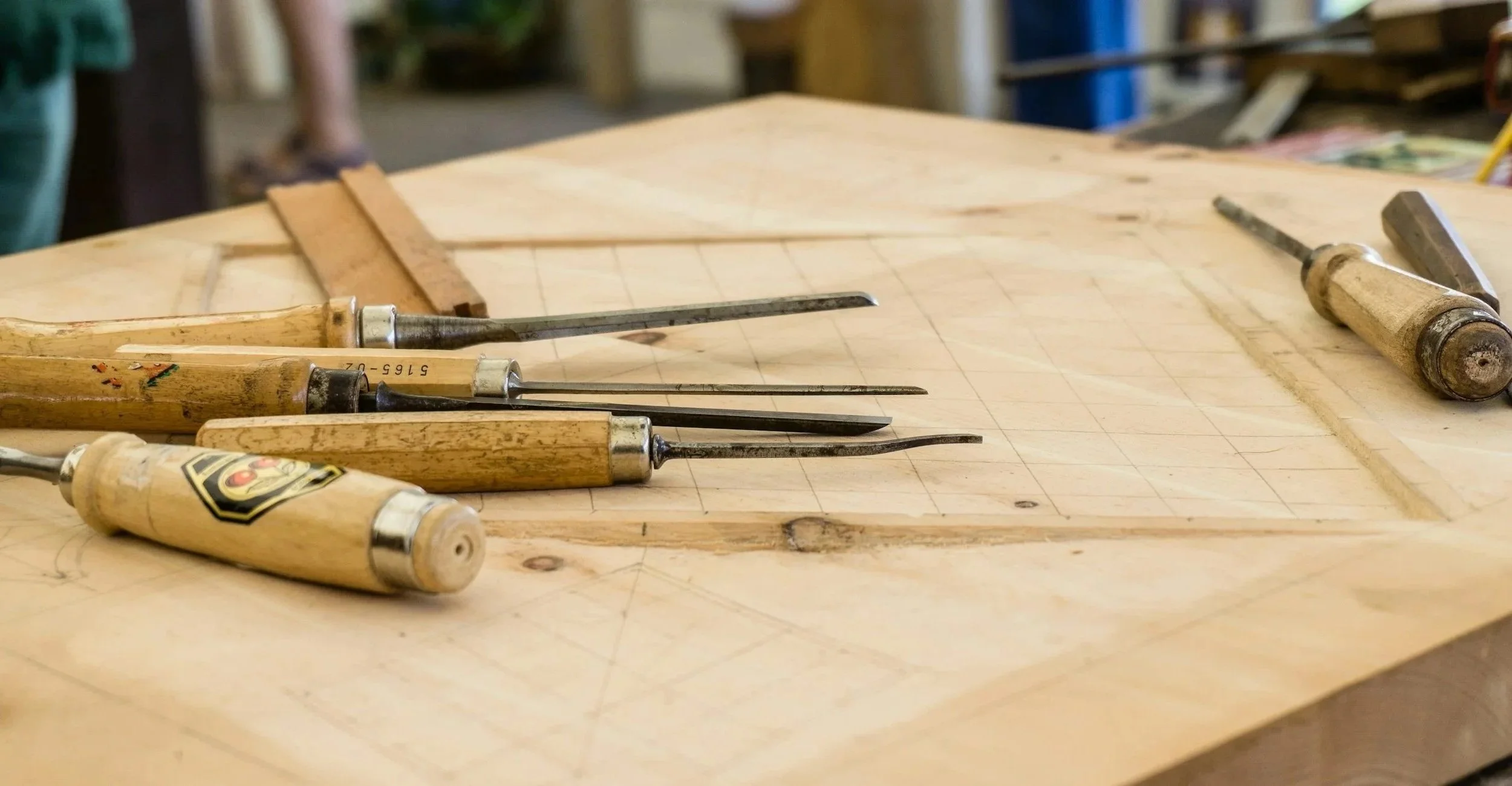 Woodworking chisels and carving tools resting on a wooden workbench with a faint grid pattern, in a woodworking shop.