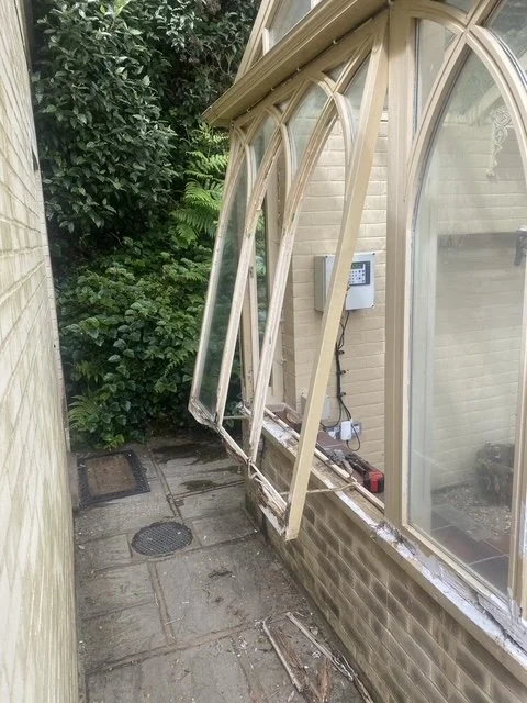 A greenhouse with a broken sliding door frame leaning against the exterior wall. The ground is paved with bricks, and there are lush green bushes nearby.