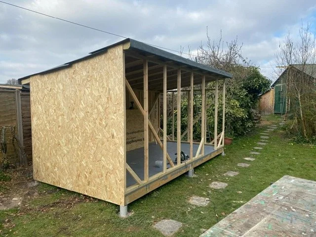 A partially constructed shed with a sloped roof, wooden frame, and plywood walls in a garden.