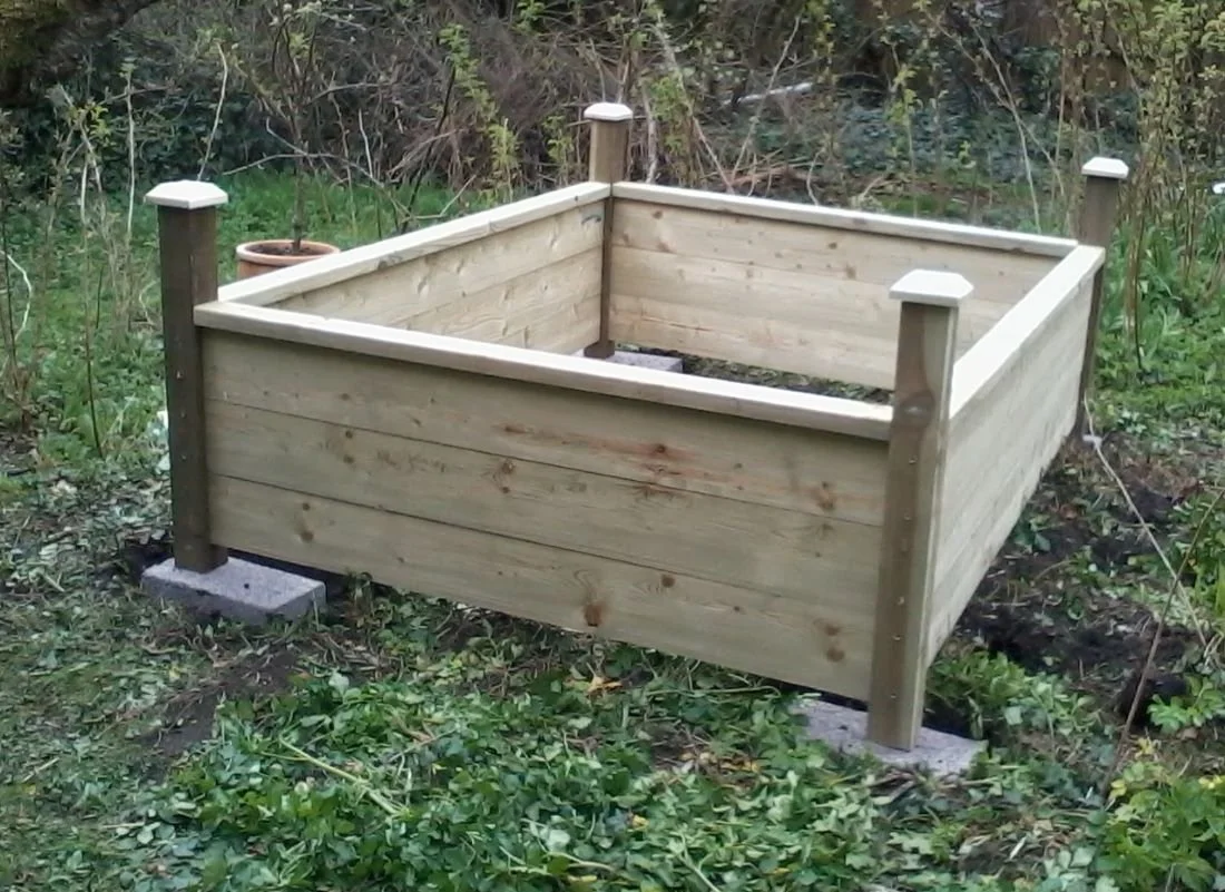 A freshly built raised wooden garden bed with four posts, situated in a garden surrounded by greenery and plants.