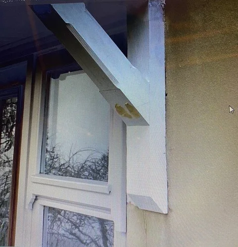 An exterior corner of a building with a white window, a white protruding structure above the window, and a white vertical block attached to the wall.