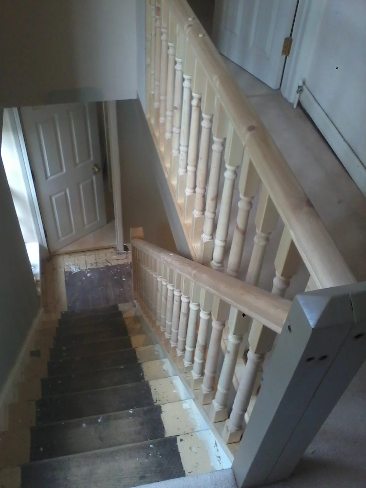 Photo of a staircase with wooden handrails and balusters, partially painted, leading down to a landing with a door and dark stairs at the bottom.