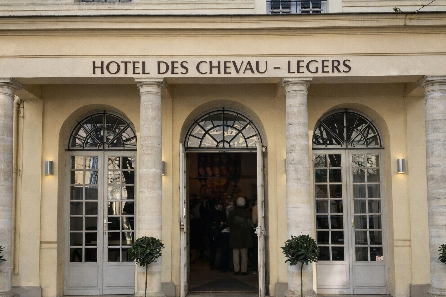 Bâtiment historique éclairé la nuit avec une façade jaune, colonnes à l'entrée, fenêtres à barres, porte métallique, et lumière extérieure, reflet des branches d'arbres sur le mur.