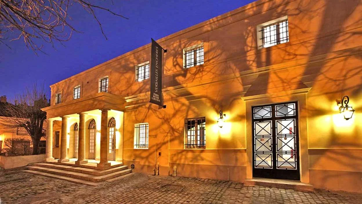 Bâtiment historique éclairé la nuit avec une façade jaune, colonnes à l'entrée, fenêtres à barres, porte métallique, et lumière extérieure, reflet des branches d'arbres sur le mur.