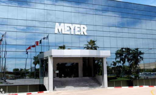 Façade d'un bâtiment avec le nom 'MEYER' affiché en gros caractères, vitres reflétant le ciel et des palmiers devant.