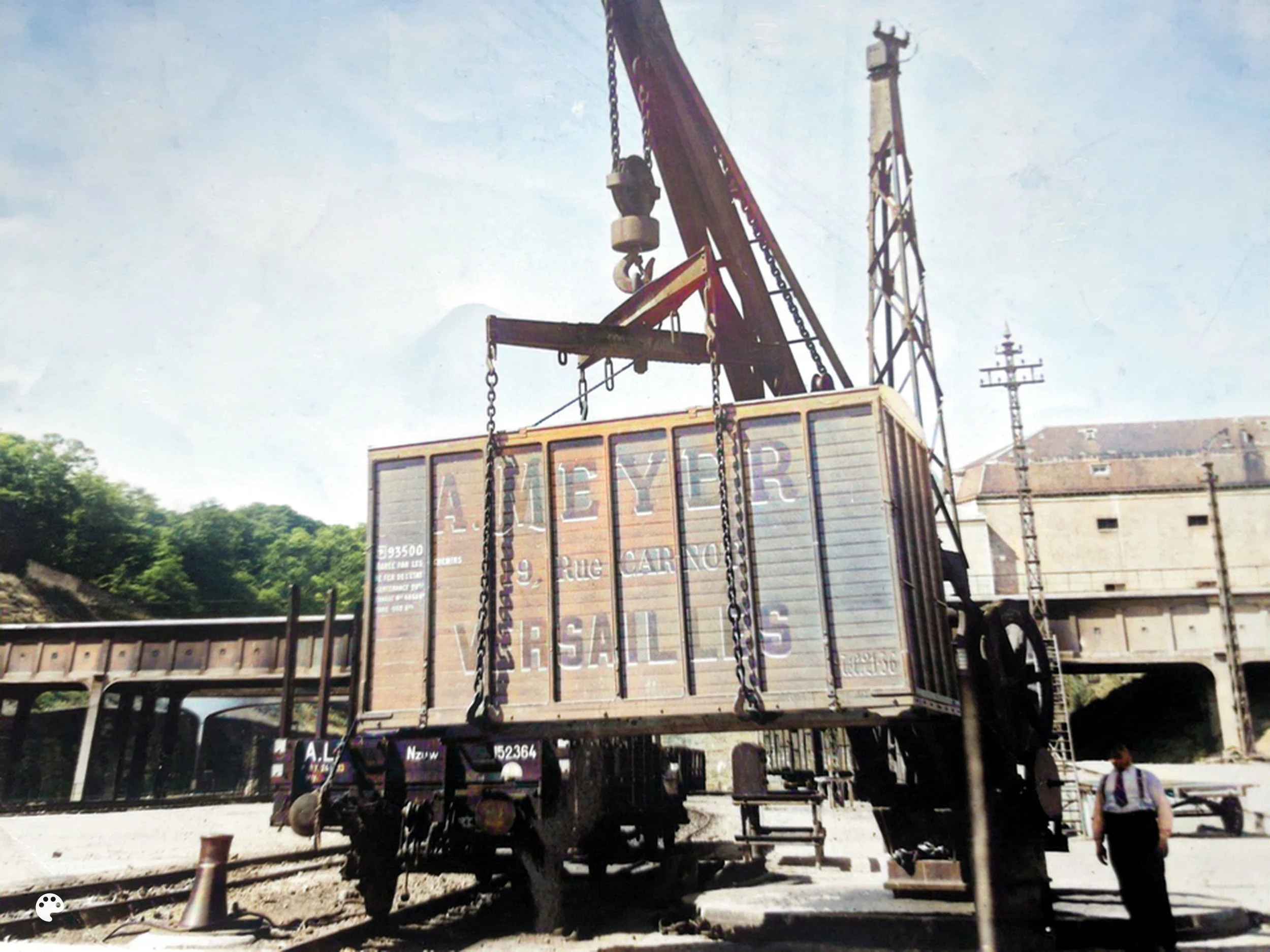 Un train de marchandises en réparation avec une grue suspendant une caisse sur les rails, un homme avec un costume blanc et une cravate à côté.