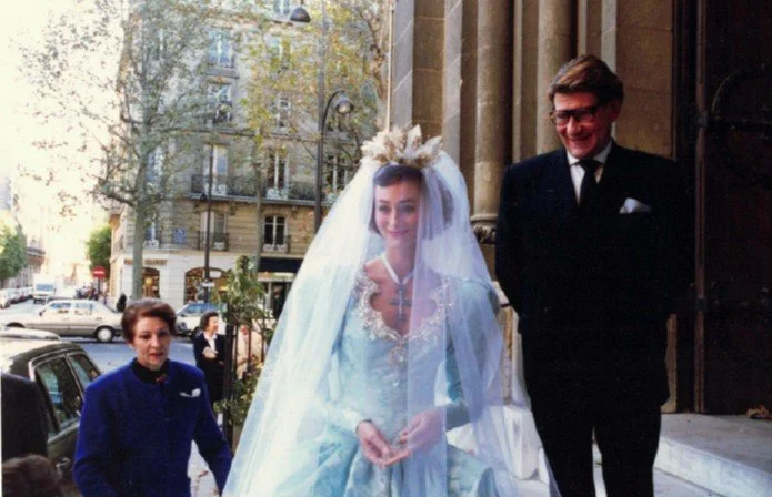 Une jeune femme en robe de mariage avec un voile et un bouquet, accompagnée de deux hommes en costumes, devant un bâtiment en pierre.