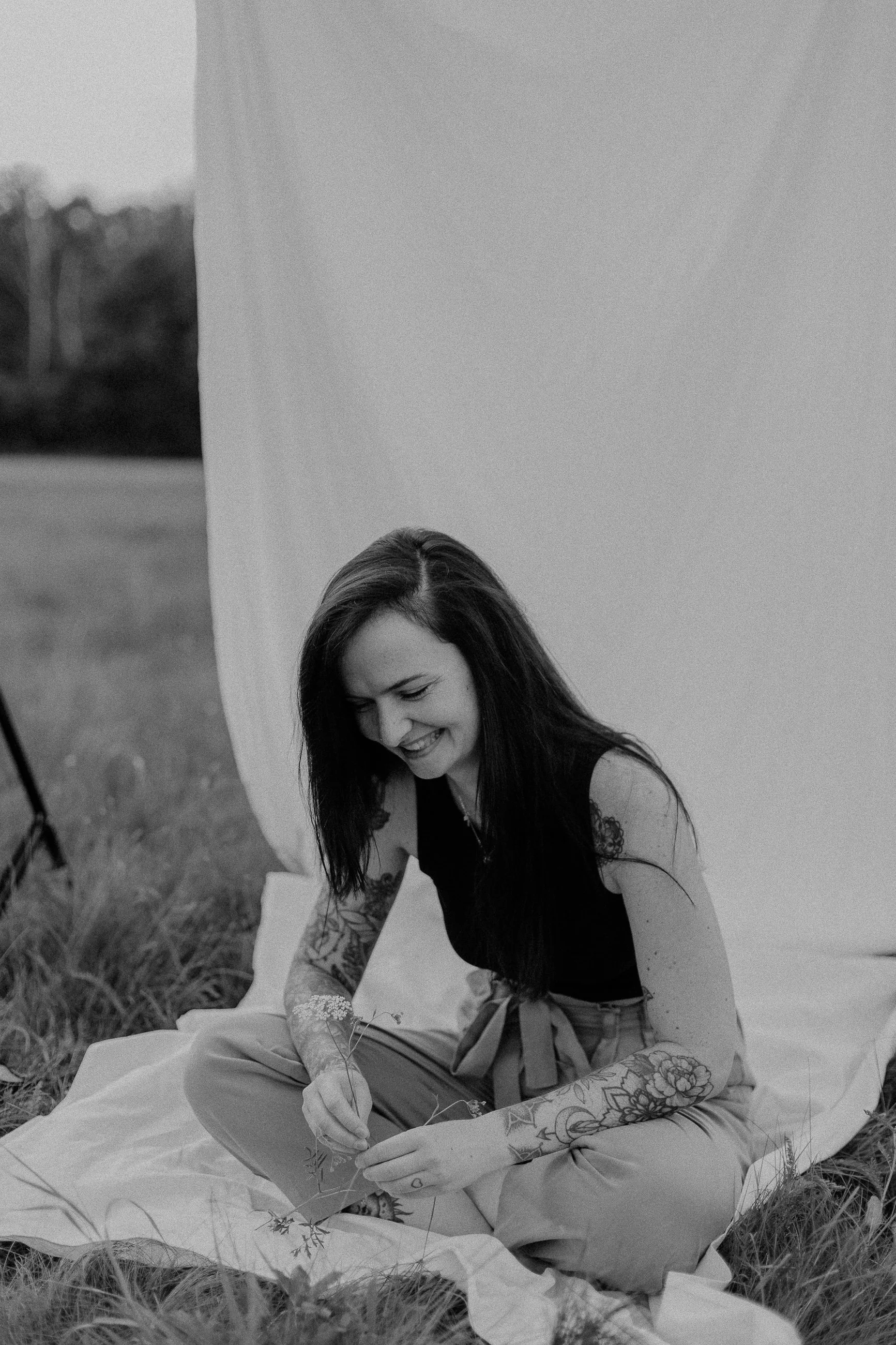 Junge Frau mit Tattoos sitzt im Freien auf einem Tuch, lächelt und hält eine kleine Pflanze in der Hand.