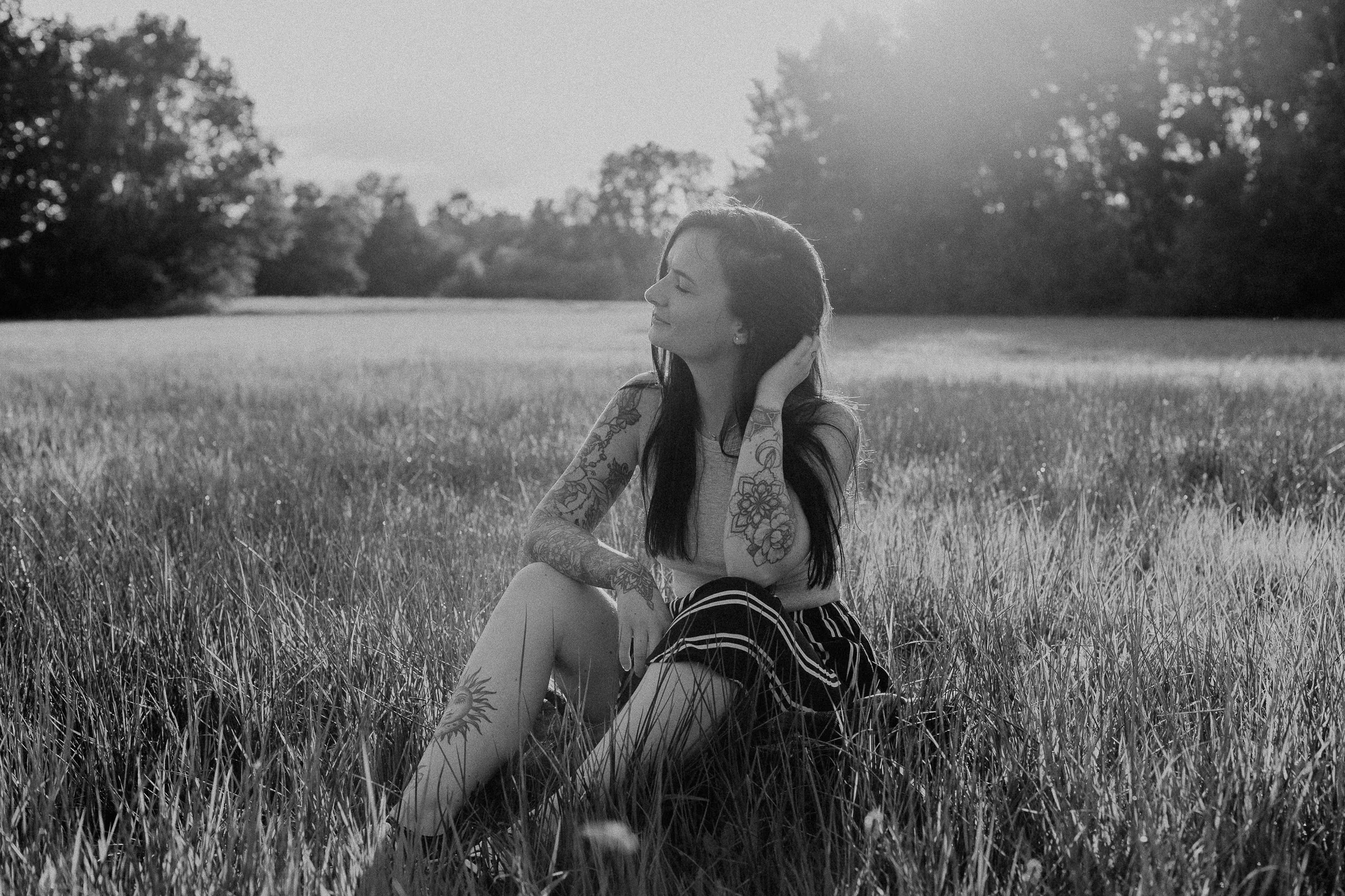 Junge Frau mit Tattoos sitzt im Gras auf einer Wiese, blickt nach oben, Sonnenlicht im Hintergrund.