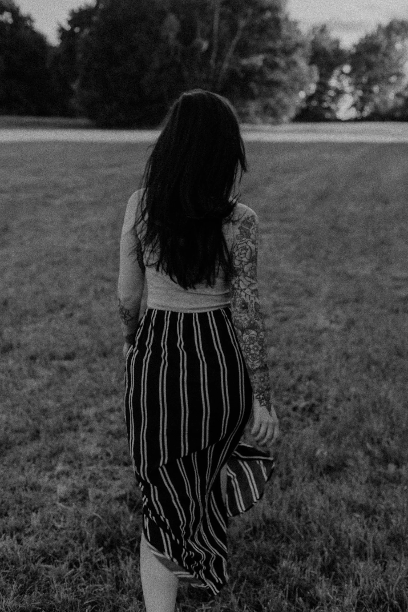Eine Frau mit dunklen Haaren, die von hinten zu sehen ist, steht auf einem offenen Rasenfeld, getragen mit gestreifter Hose und einem Top, im Hintergrund Bäume.