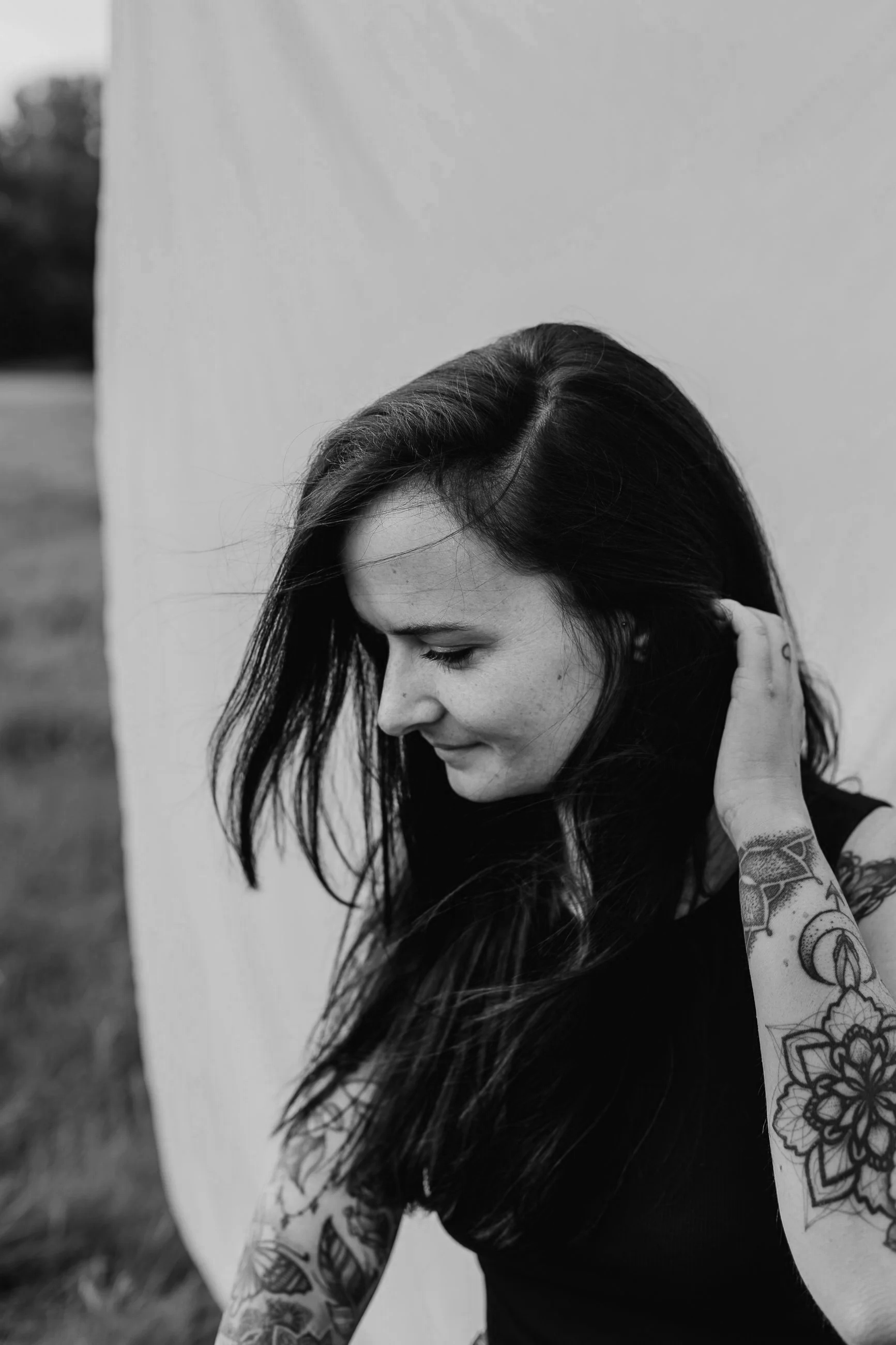 Schwarz-weiß-Foto einer jungen Frau mit langen dunklen Haaren, die lächelnd den Kopf neigt und an ihrem Hals kratzen, mit sichtbaren Tattoos auf Armen, vor einem weißen Hintergrund, im Freien.