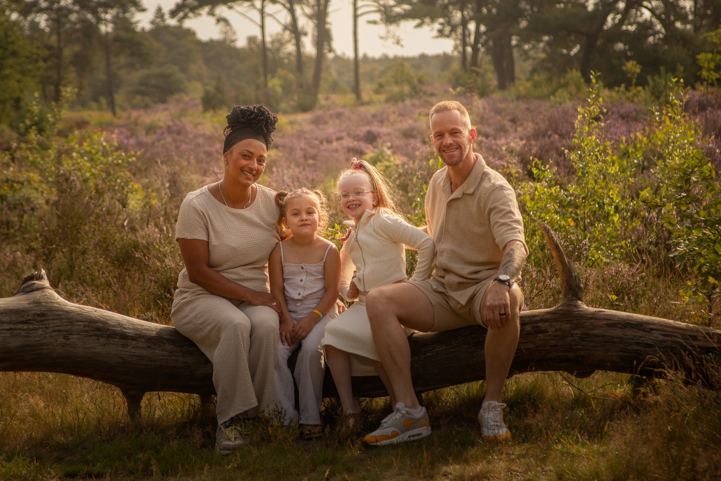 Family of four sitting on a fallen tree in a scenic outdoor setting with purple heather and trees in the background, smiling at the camera.