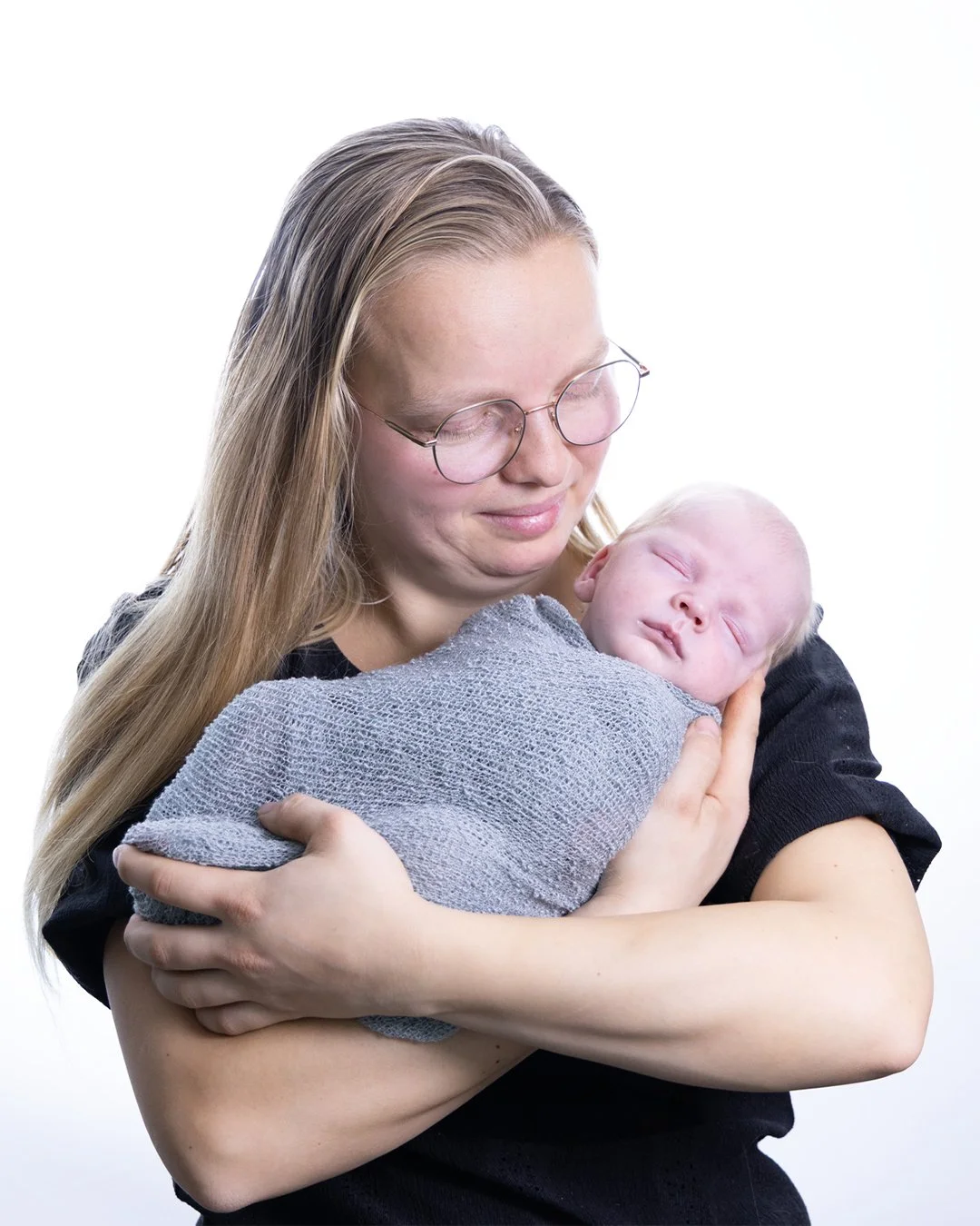 A woman with long blonde hair and glasses holding a sleeping baby wrapped in a gray blanket against a white background.