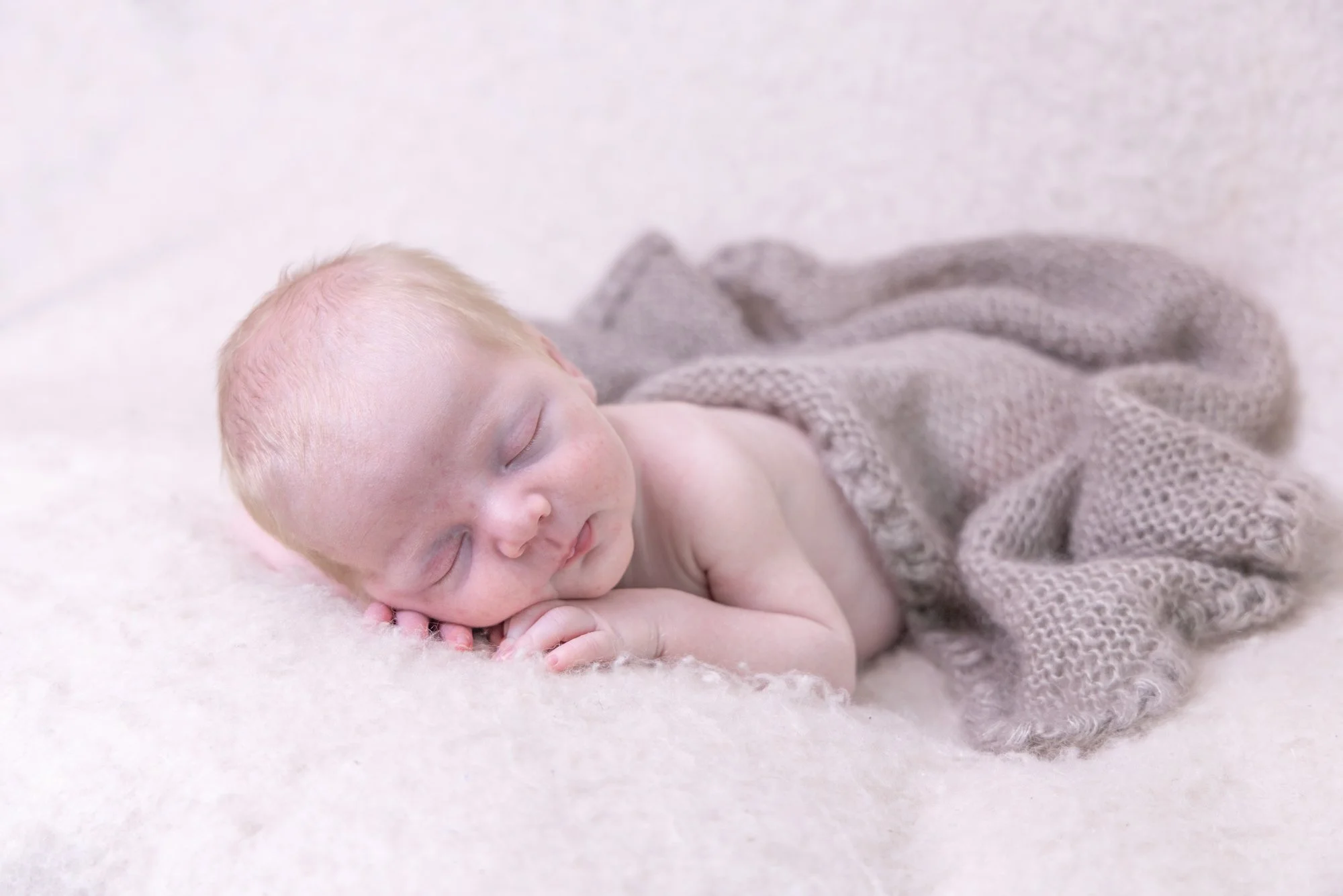 A sleeping baby with blonde hair lying on a soft, cream-colored blanket, partially covered by a gray knitted blanket.