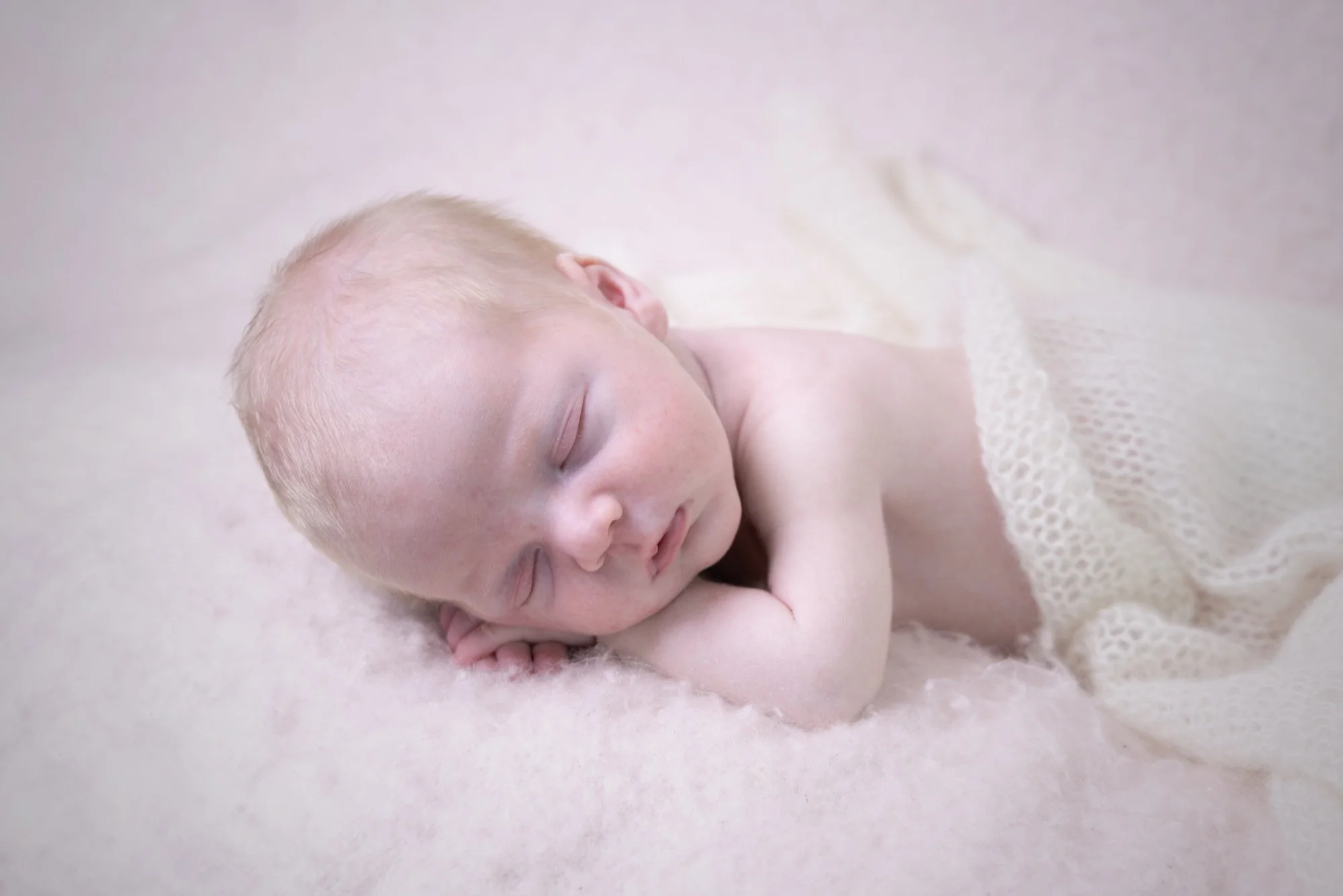 A sleeping newborn baby with blonde hair curled on a soft pink surface, partially covered with a white textured blanket.