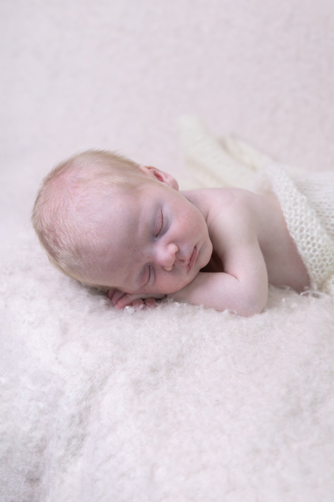 A sleeping baby with fair skin and light hair, lying on its side on a soft, fluffy surface, covered with a light-colored knitted cloth.