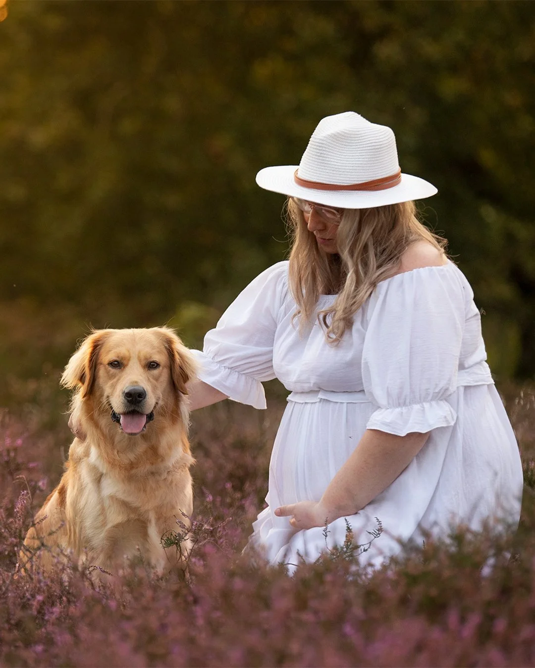 A pregnant woman wearing a white dress and a wide-brimmed hat kneeling in a field of purple flowers with a golden retriever sitting next to her.