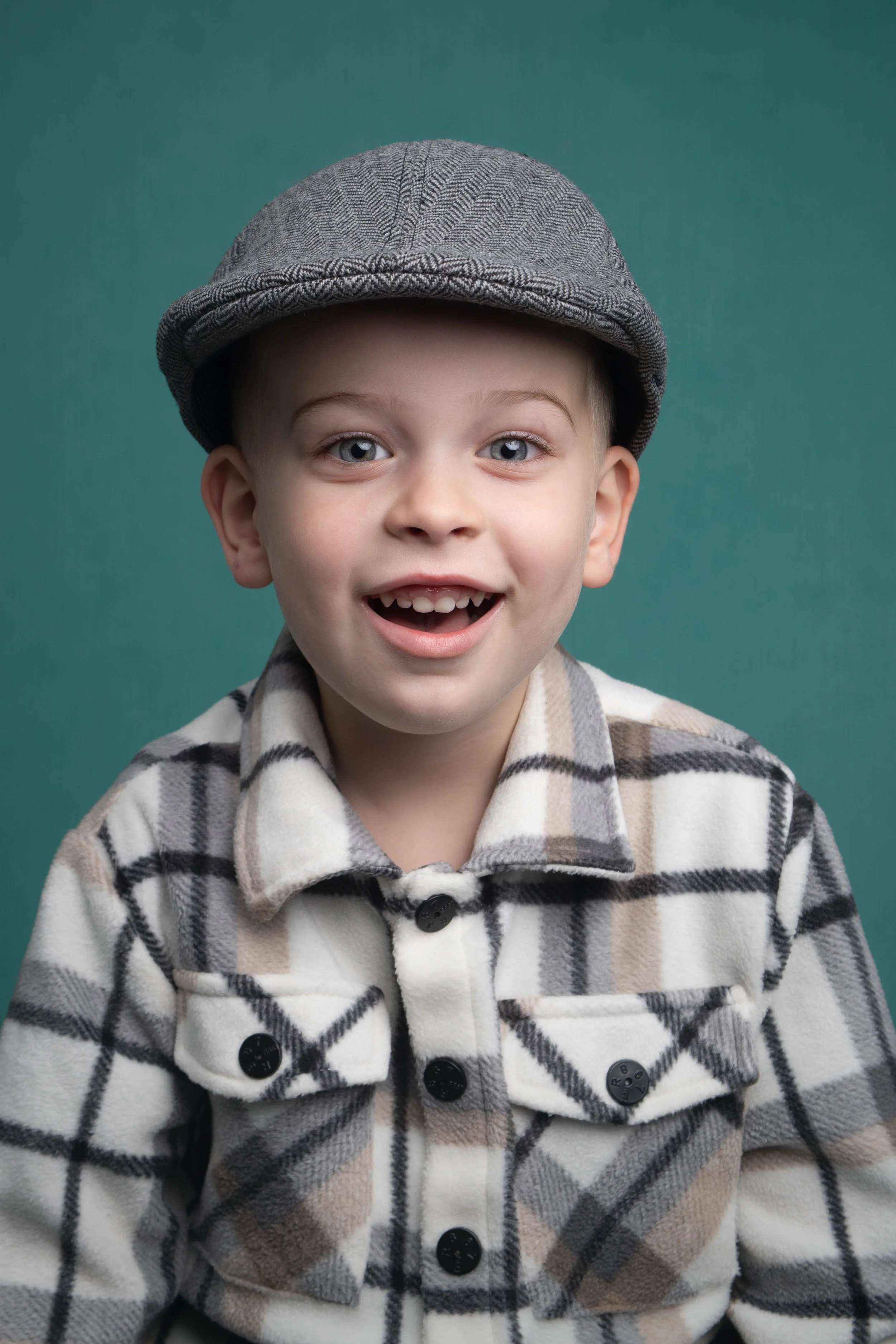 A young boy smiling wearing a gray plaid newsboy cap and a matching plaid jacket, against a solid teal background.