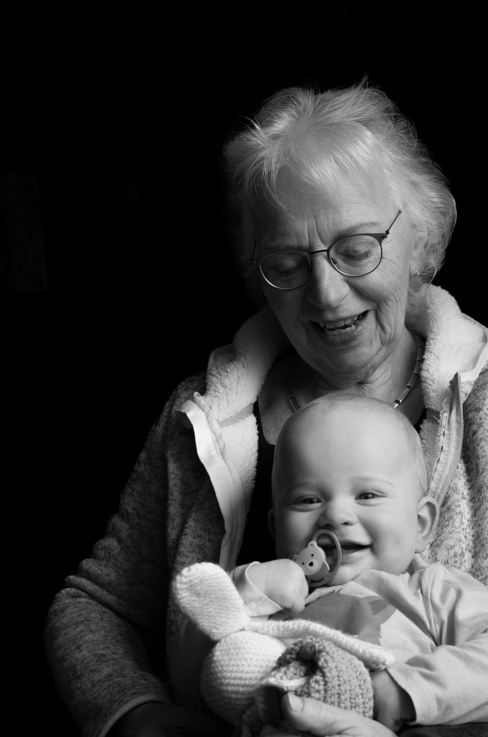 An elderly woman with glasses holding a smiling baby with a pacifier against a dark background.
