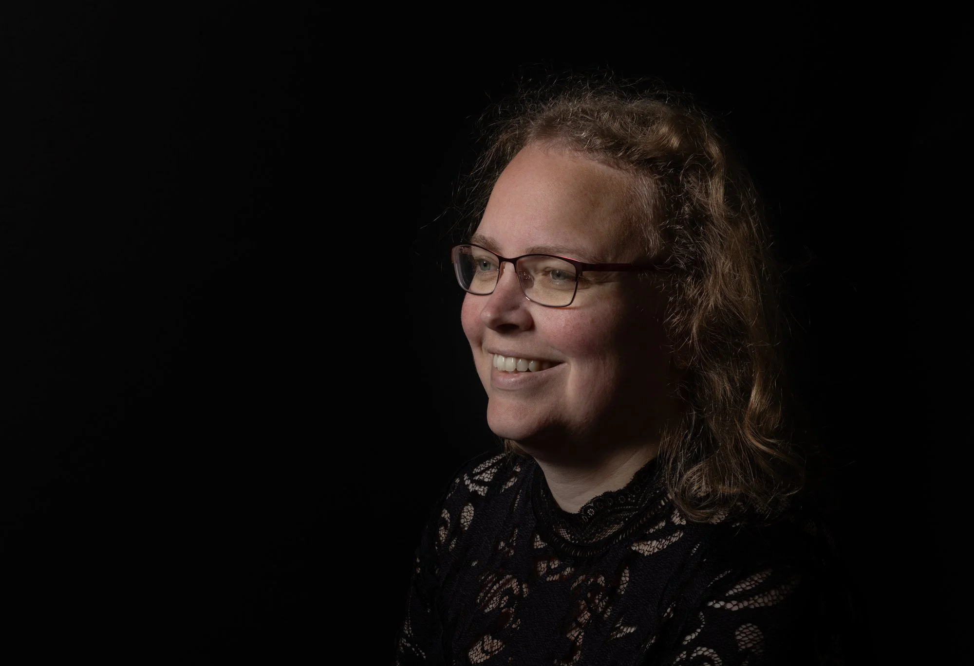 A woman with glasses and curly hair smiling, wearing a black lace top, against a black background.