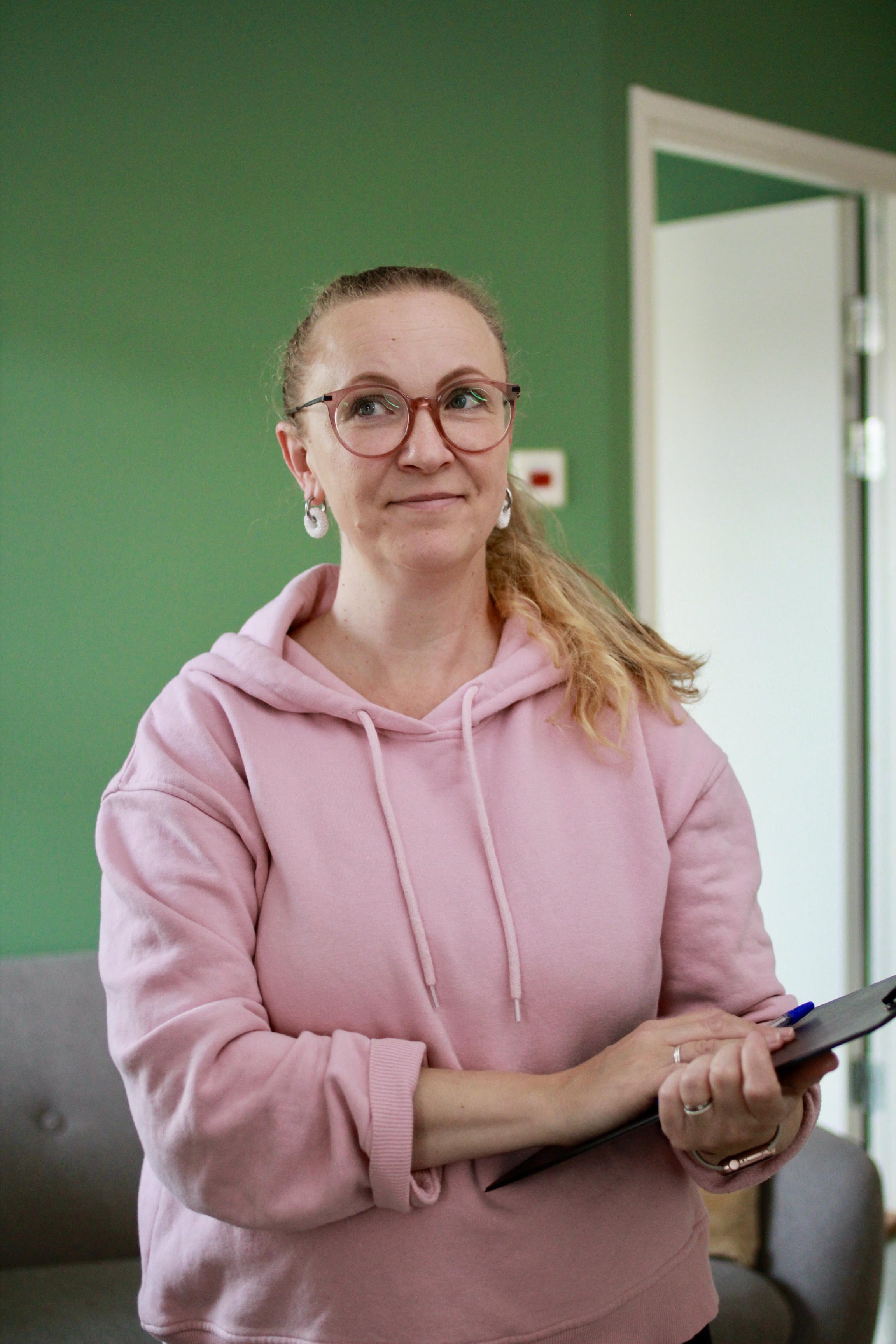 Nainen hymyilee, hänellä on punasävyiset kehykset ja hän pitää tablet-laitetta kädessään, taustalla vihreä seinä ja avoin ovi.