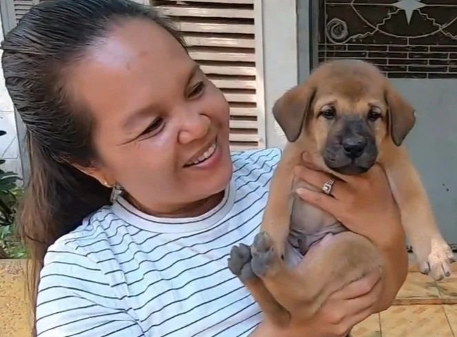 Kong smiling and holding a light brown puppy at a pagoda in Siem Reap who she fed, with darker markings on its face outside near a door.