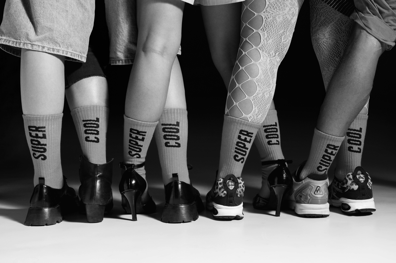Close-up of the lower legs and feet of five women standing side by side, wearing socks with the words 'SUPERCOOL', and various shoes including high heels, sneakers, and boots, on a dark background.