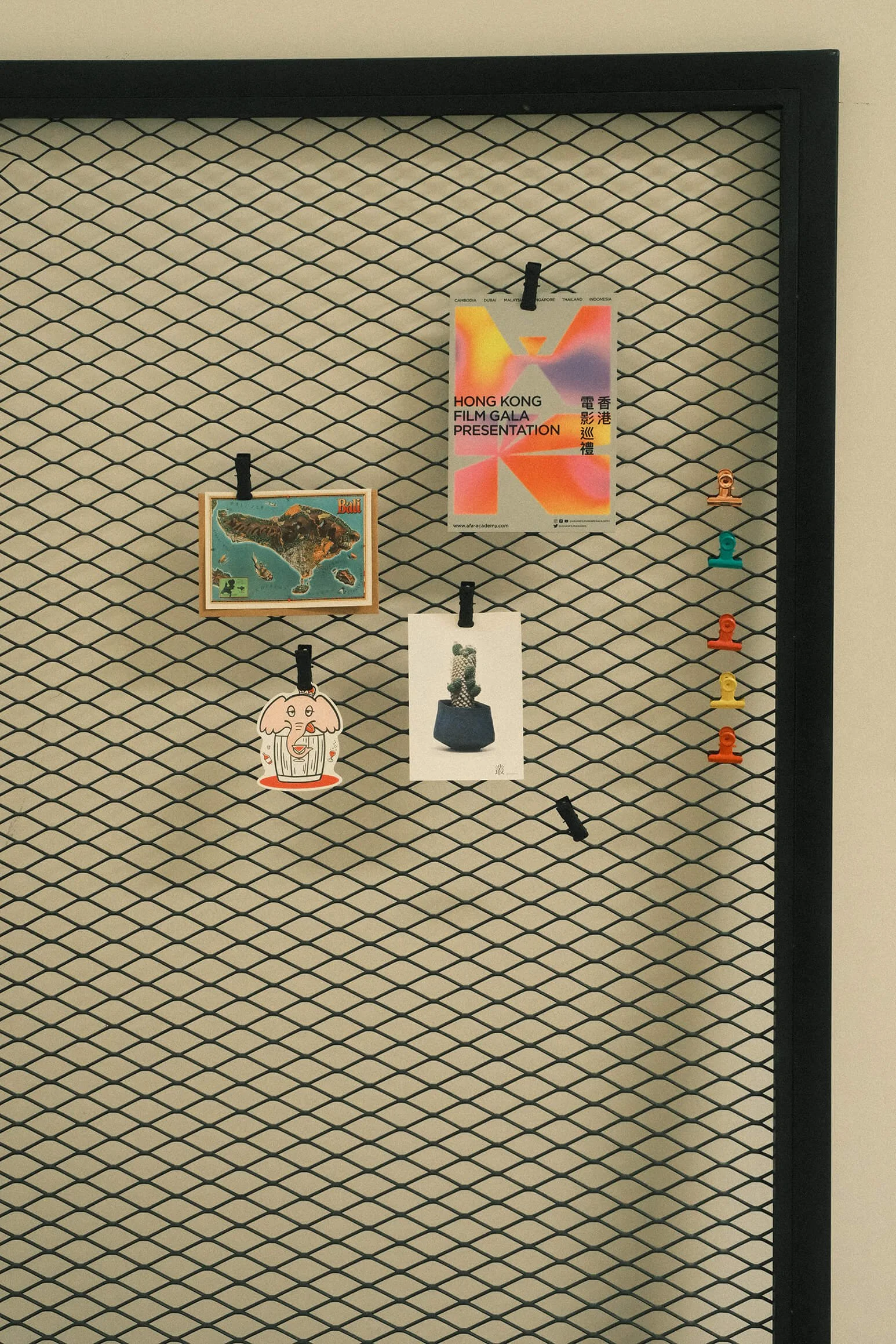 Inside the Supercool Studio Bali. A black wire mesh bulletin board with various photos, posters, and clips attached, including a map of Bali, an illustration of a cute character, and promotional materials for the Hong Kong Film Gala.