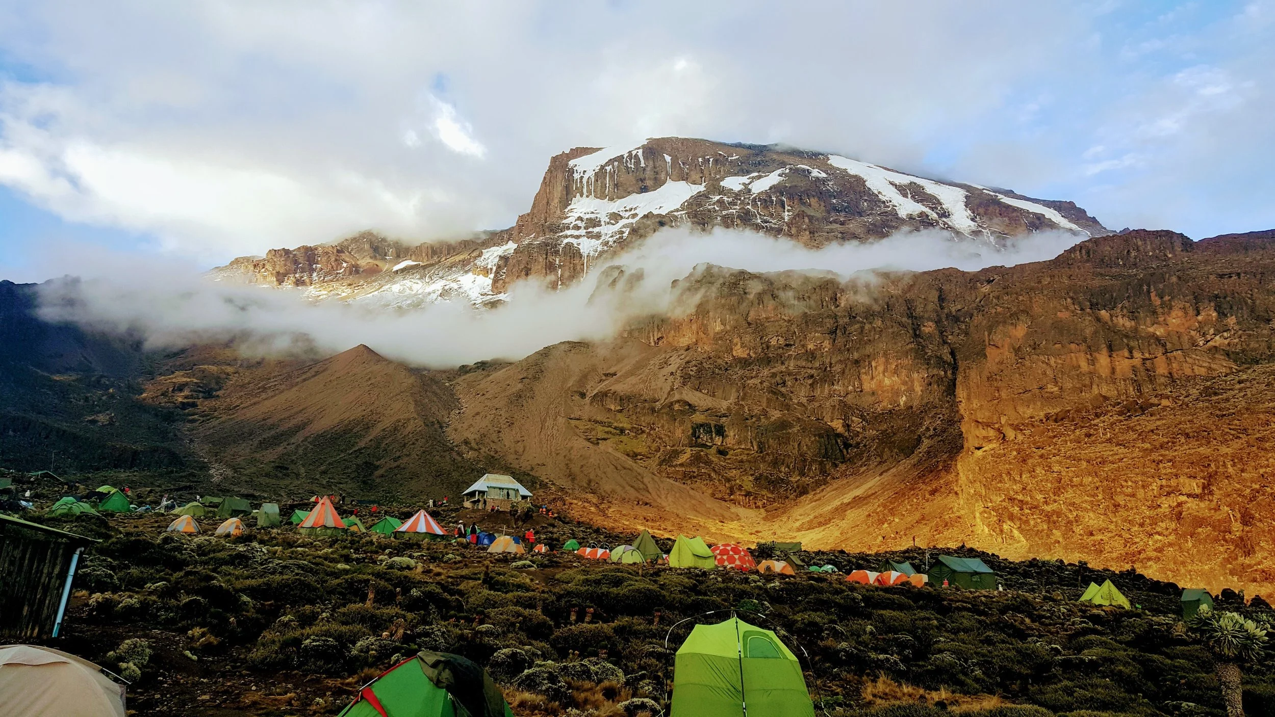 Kampeerplaatsen op een camping in een bergachtig gebied met tenten en een bergtop met sneeuw en wolken eromheen.