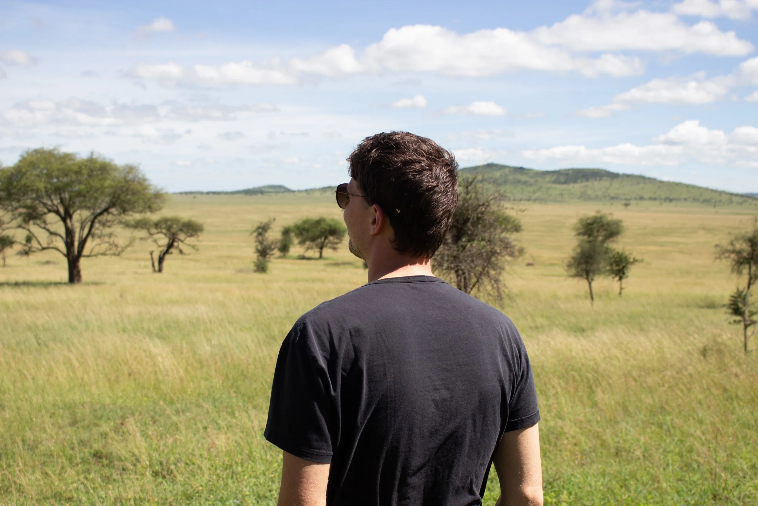 Man met donkere haar en een zwarte T-shirt kijkt uit over een grasvlakte met enkele bomen, onder een blauwe hemel met witte wolken.