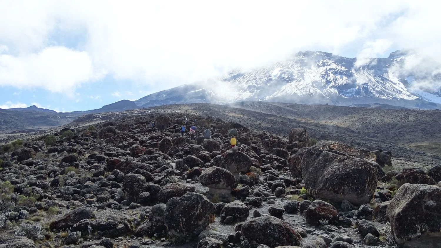 Wandelaars in een rotsachtige vlakte met op de achtergrond een besneeuwde bergtop, tijdens de beklimming van de Kilimanjaro via de Machame-route.