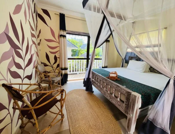 Kamer met een wit bed met een klamboe, twee houten stoelen, een ronde natuurkleurige tapijt, een deur naar een balkon met uitzicht op bomen en een huis, en bloemenbehang op een muur.