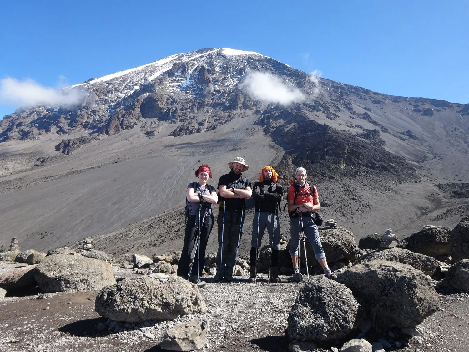 Vier mensen op een berghelling bij de beklimming van de Kilimanjaro, gekleed in outdoors kleding en met rugzakken en wandelstokken, onder een heldere blauwe hemel.
