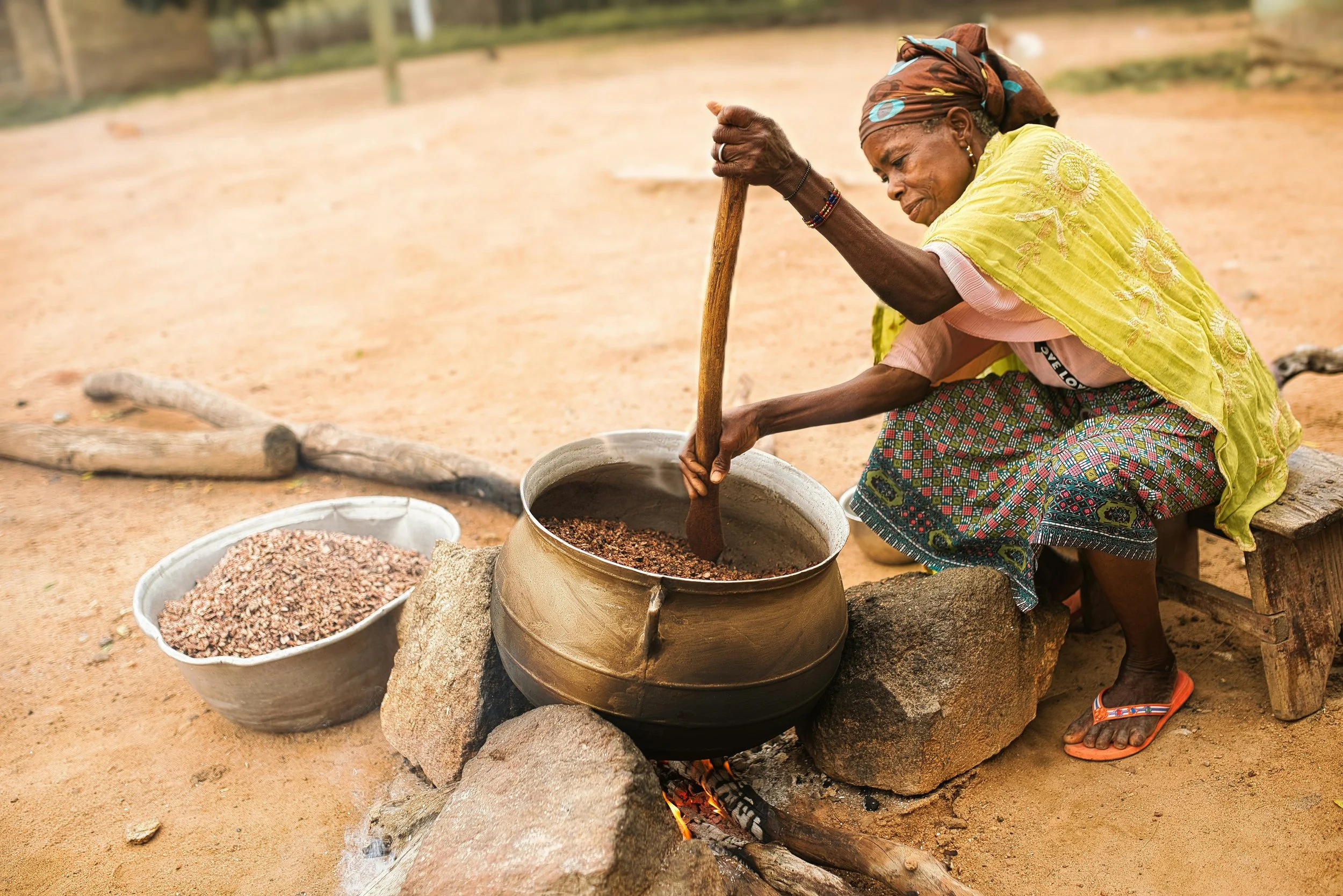 Een oudere vrouw, gekleed in kleurrijke kleding en een hoofddoek, roert met een houten lepel in een grote gietijzeren pot boven een open vuur in een eenvoudige buitenkeuken. Een metalen schaal met zaden of granen ligt ernaast.