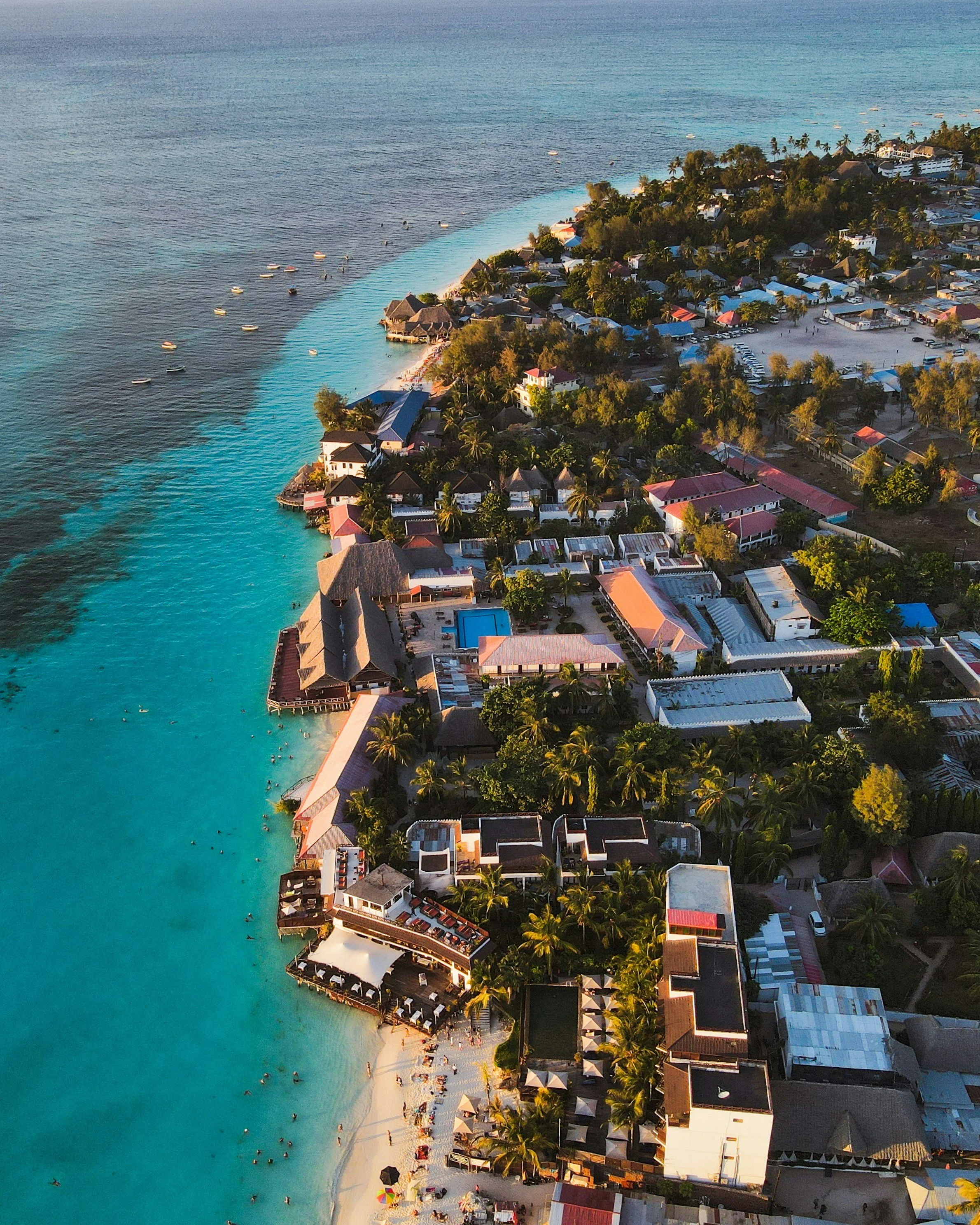Luchtfoto van een tropisch strandresort met blauwe zee, wit zandstrand, ligstoelen, parasols, en veel palmbomen, gebouwen en bungalowtenten.