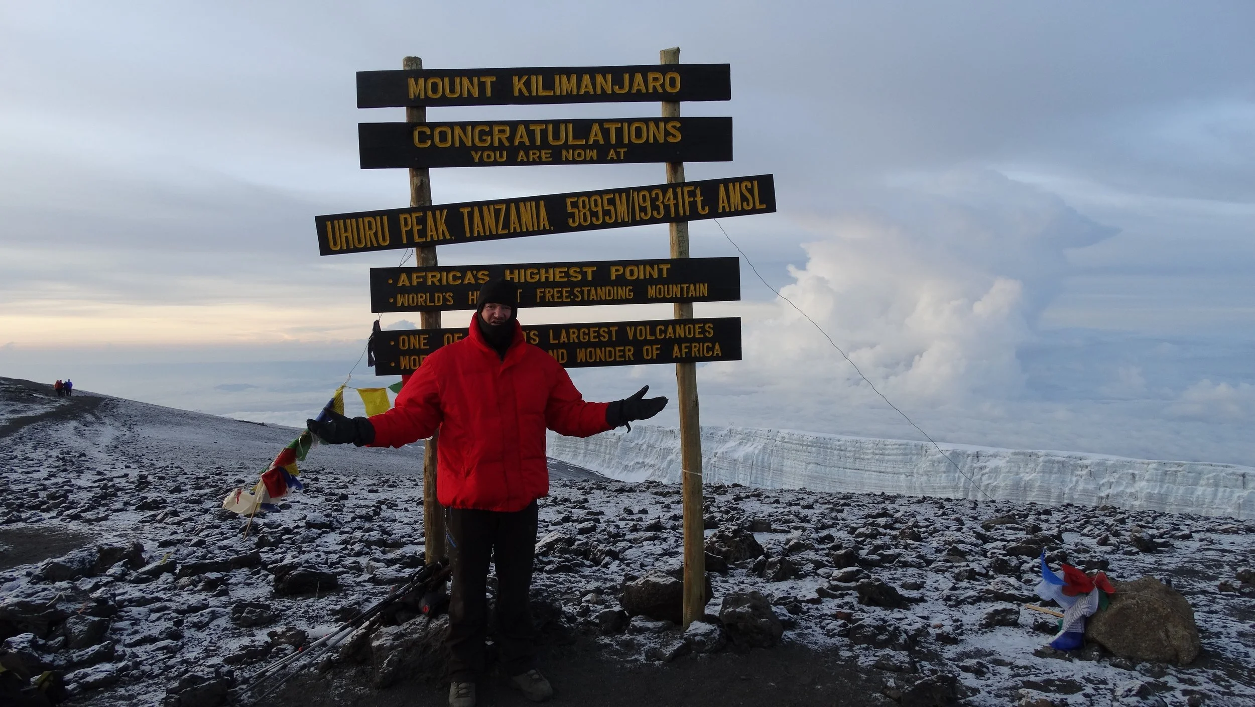 Persoon in rode jas bij het bord van de Uhuru Peak op Mount Kilimanjaro in Tanzania, met een achtergrond van een gletsjer en een bewolkte hemel.