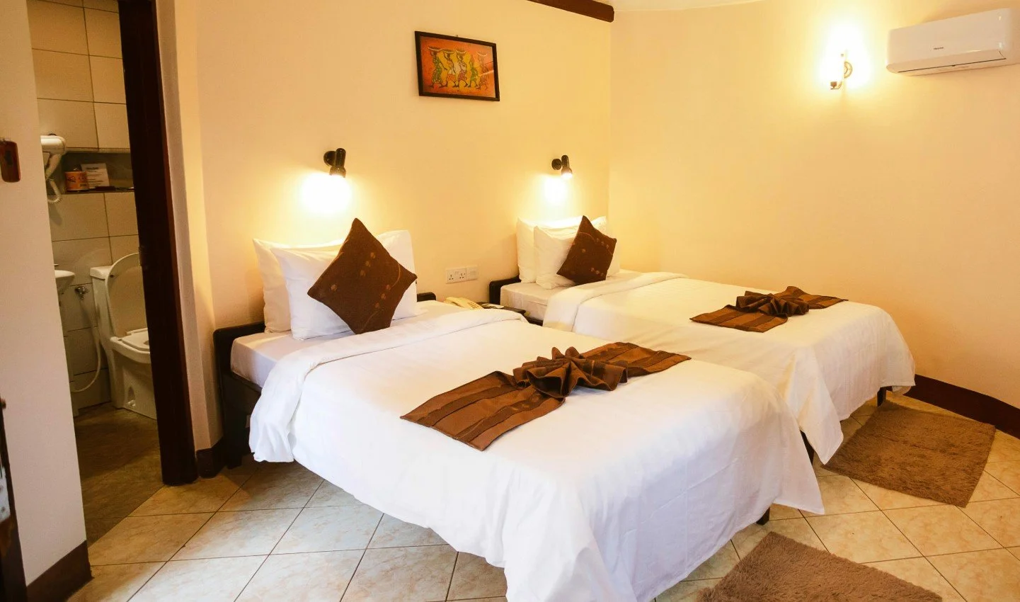 Een hotelkamer met twee eenpersoonsbedden, elk met een gekruiste lakentje, wit beddengoed en een bruin kussen, lampen aan de muur en een airconditioner.