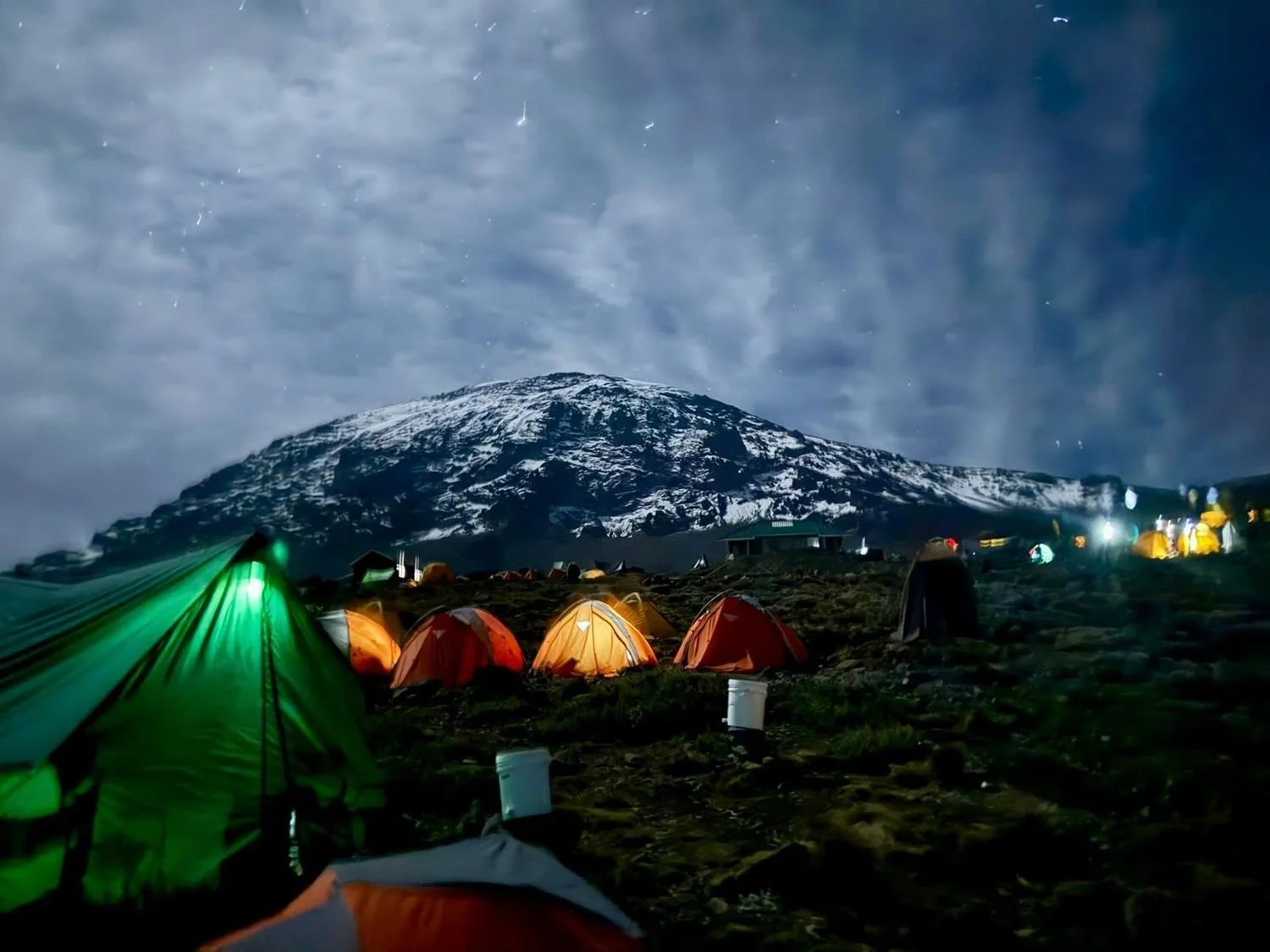 Tentenkamp onder een bewolkte nachtelijke sky met een besneeuwde berg op de achtergrond.
