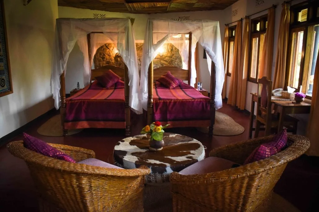 Een gezellige hotelkamer met twee bedden met netgemaakte witte doeken eromheen, een zithoek met twee wicker stoelen en een ronde tafel met bloemen, grote ramen met gouden gordijnen en een werktafel met een telefoon en bloemen.