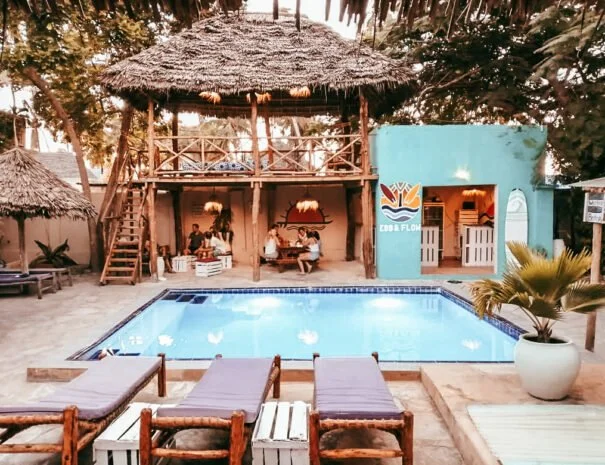 Tropisch zwembad met houten terras en zonbedden, een gebouw met blauw muur en een prieel boven het zwembad, omgeven door palmbomen en bomen.