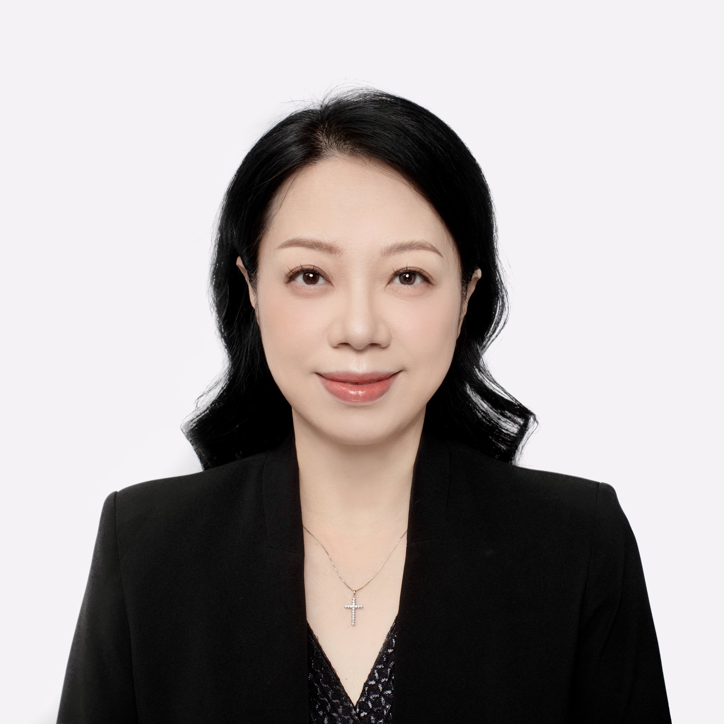 A woman with black hair in soft waves, wearing a black blazer and a silver cross necklace, smiling against a plain white background.