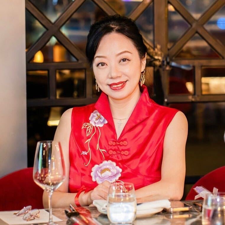 A woman wearing a red traditional Asian dress with embroidered flowers, sitting at a dining table with wine and water glasses.