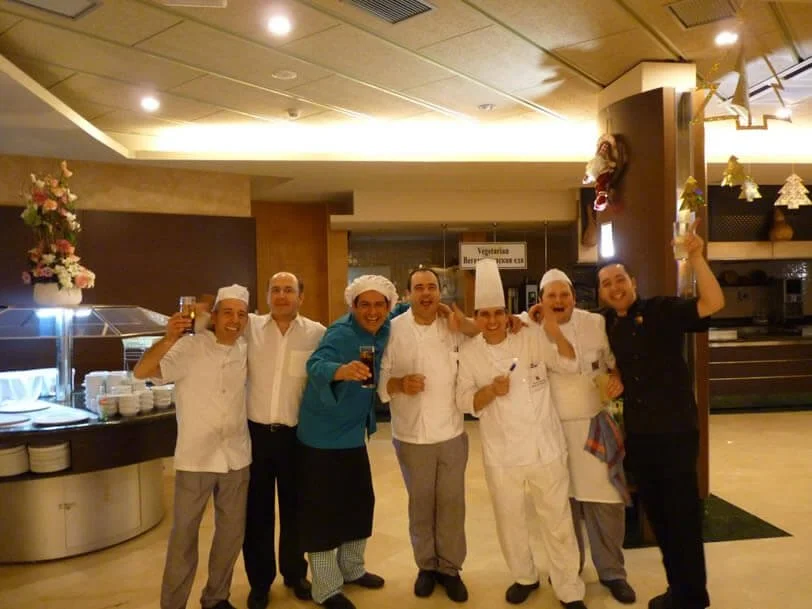 Group of seven chefs and staff celebrating with drinks in a restaurant kitchen or dining area.