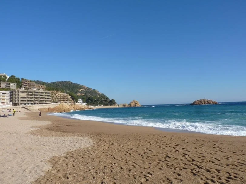 View of a sandy beach with a few people, ocean waves, rocky formations, and buildings on a hillside under a clear blue sky.