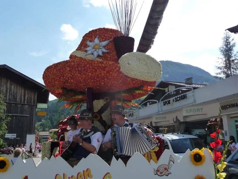 A parade float decorated with orange and yellow flowers, shaped like a large hat with a white and blue flower, with musicians playing instruments in front of it, in a mountain town square.