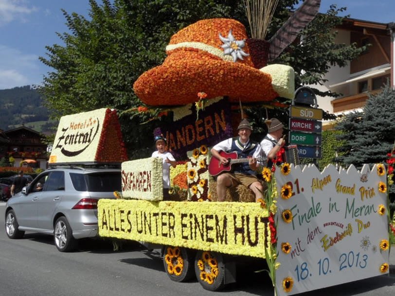 A parade float decorated with flowers, featuring a large mushroom hat, a musician playing guitar, and children dressed in traditional clothes, with a sign celebrating a festival in Meran on October 18, 2013.
