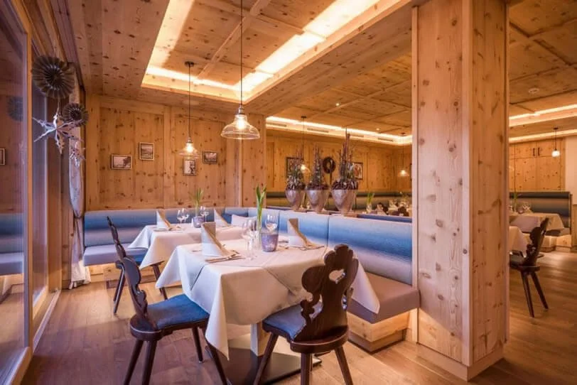 A cozy dining area with wooden walls and ceiling, decorated with framed pictures and ornaments, featuring a table set with white napkins, glasses, and a centerpiece, surrounded by a mix of traditional and modern chairs.